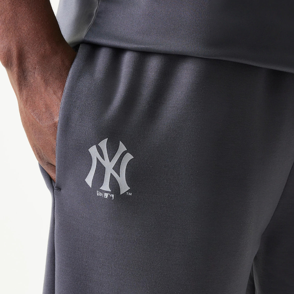 New Era New York Yankees MLB Dark Grey Joggers, close-up view showing the white New York Yankees logo printed on the side of the joggers, with a fabric texture and a hand resting in the side pocket.