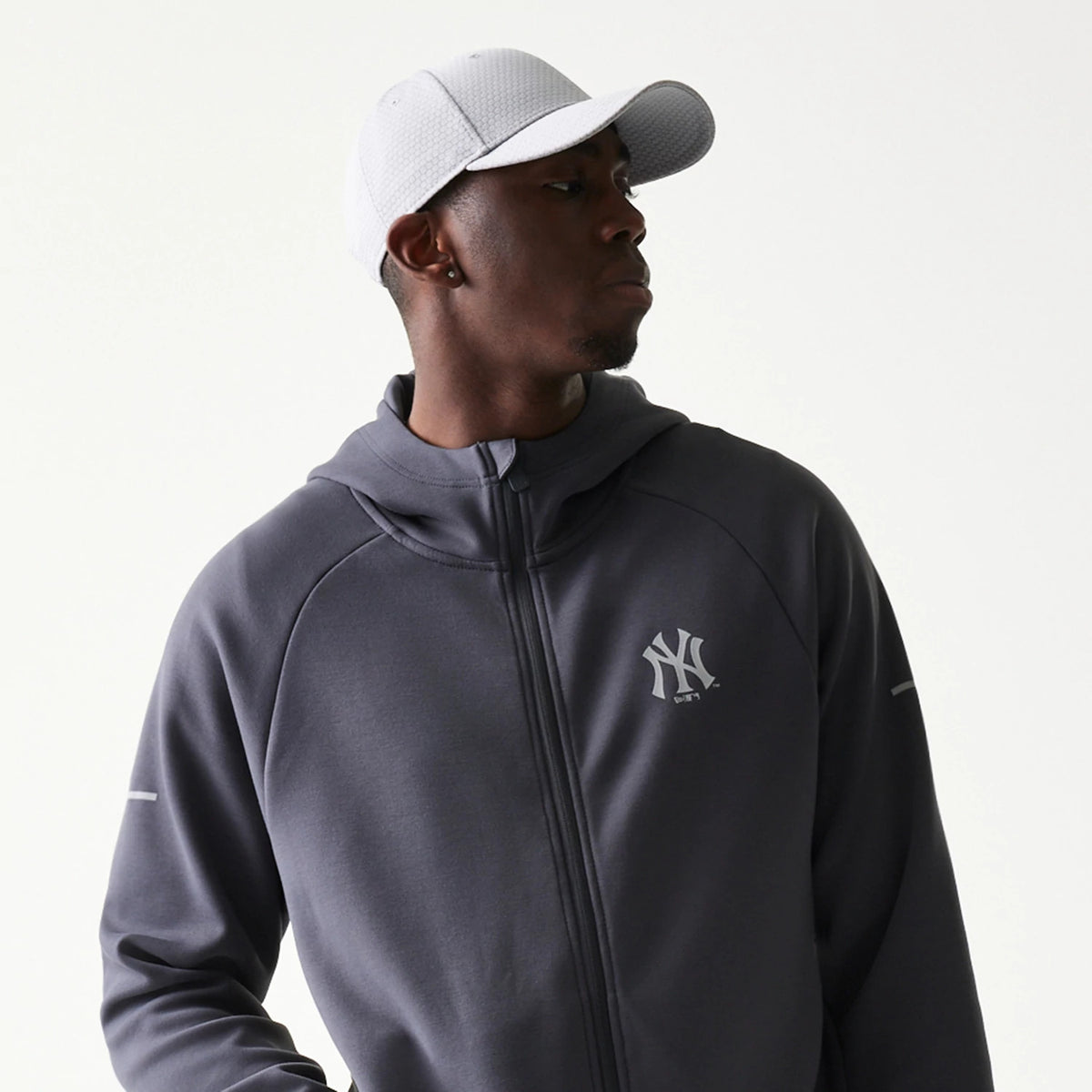 New Era New York Yankees MLB Dark Grey Full Zip Hoodie, front three-quarter view showing a grey hoodie with a full zip and an embroidered Yankees logo on the left chest, worn by a model also styling a plain white cap.