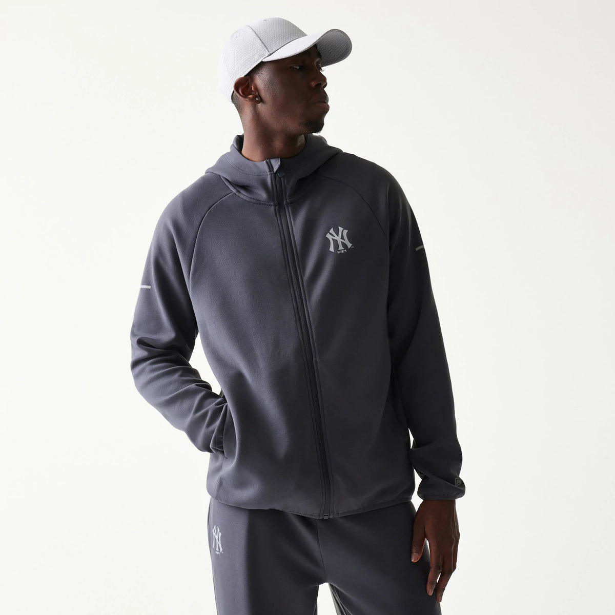 New Era New York Yankees MLB Dark Grey Full Zip Hoodie, front view on-model displaying the hoodie zipped up and featuring a white embroidered Yankees logo on the left chest, with matching tracksuit bottoms and a white cap.