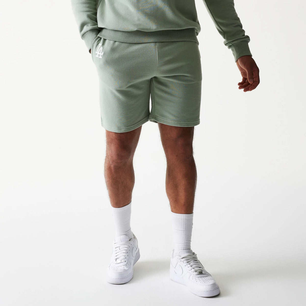 New Era LA Dodgers MLB League Essential Midi Green Shorts, front view on-model displaying a light green hue with an embroidered white LA logo on the left thigh, paired with a matching sweatshirt and white trainers.