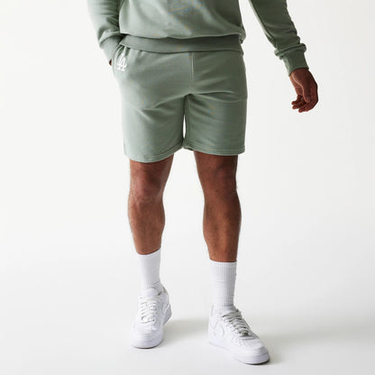 New Era LA Dodgers MLB League Essential Midi Green Shorts, front view on-model displaying a light green hue with an embroidered white LA logo on the left thigh, paired with a matching sweatshirt and white trainers.