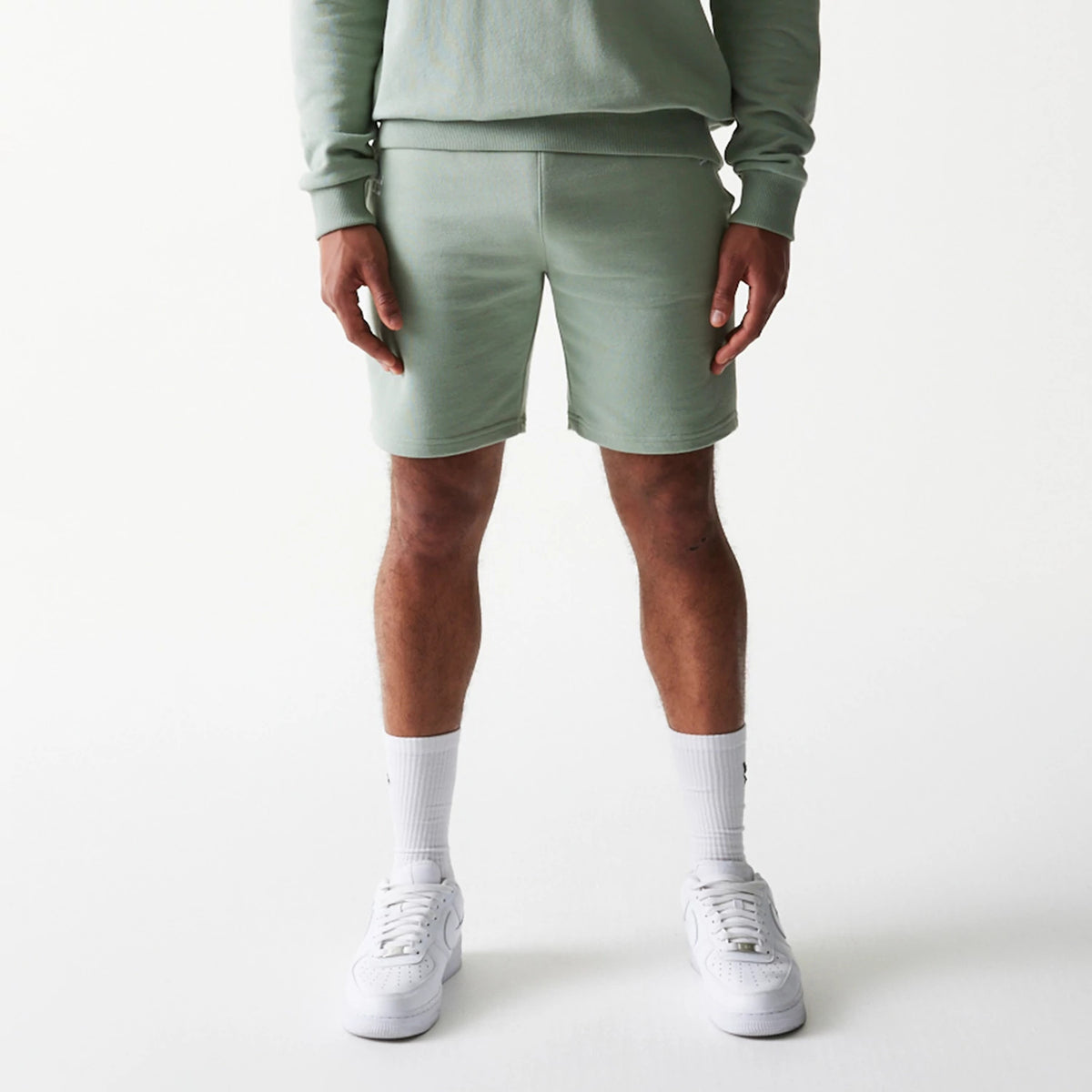 New Era LA Dodgers MLB League Essential Midi Green Shorts, front view on a model showing the shorts in a soft green with a mid-thigh length, styled with a matching green top and white trainers for a coordinated look.