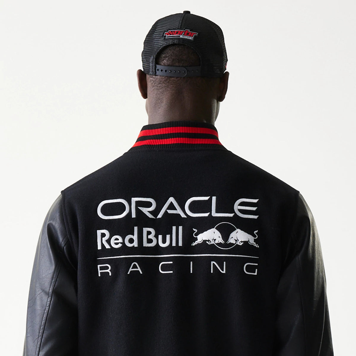 New Era Red Bull Racing Black Varsity Jacket, back view showing the large printed Oracle Red Bull Racing logo across the shoulders, with black sleeves and a red striped collar, worn by a model wearing a black cap.
