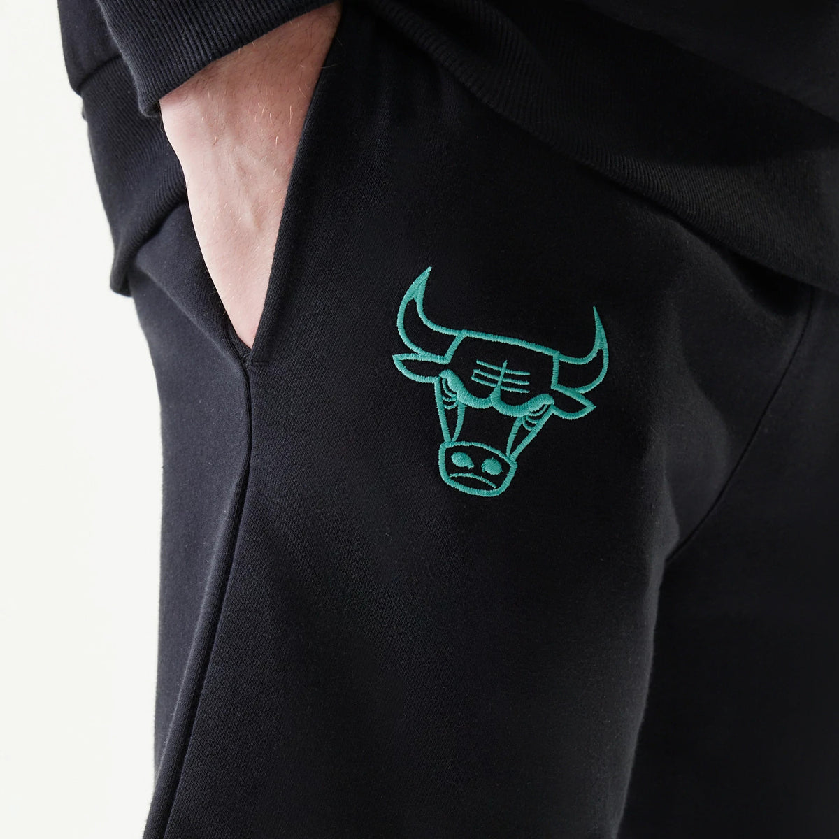 New Era Chicago Bulls NBA Core Black Shorts, three-quarter view showing a hand tucked into the side pocket and a green embroidered Chicago Bulls logo above the knee on the plain black fabric.