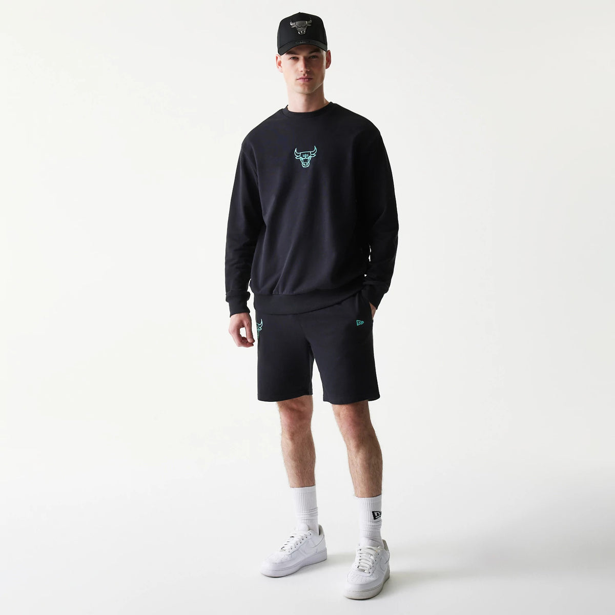 New Era Chicago Bulls NBA Core Black Shorts, front view on-model showing an embroidered teal bulls logo on the left leg, complemented by a matching sweatshirt, New Era logo on the right leg, and a cap styled for a cohesive look.