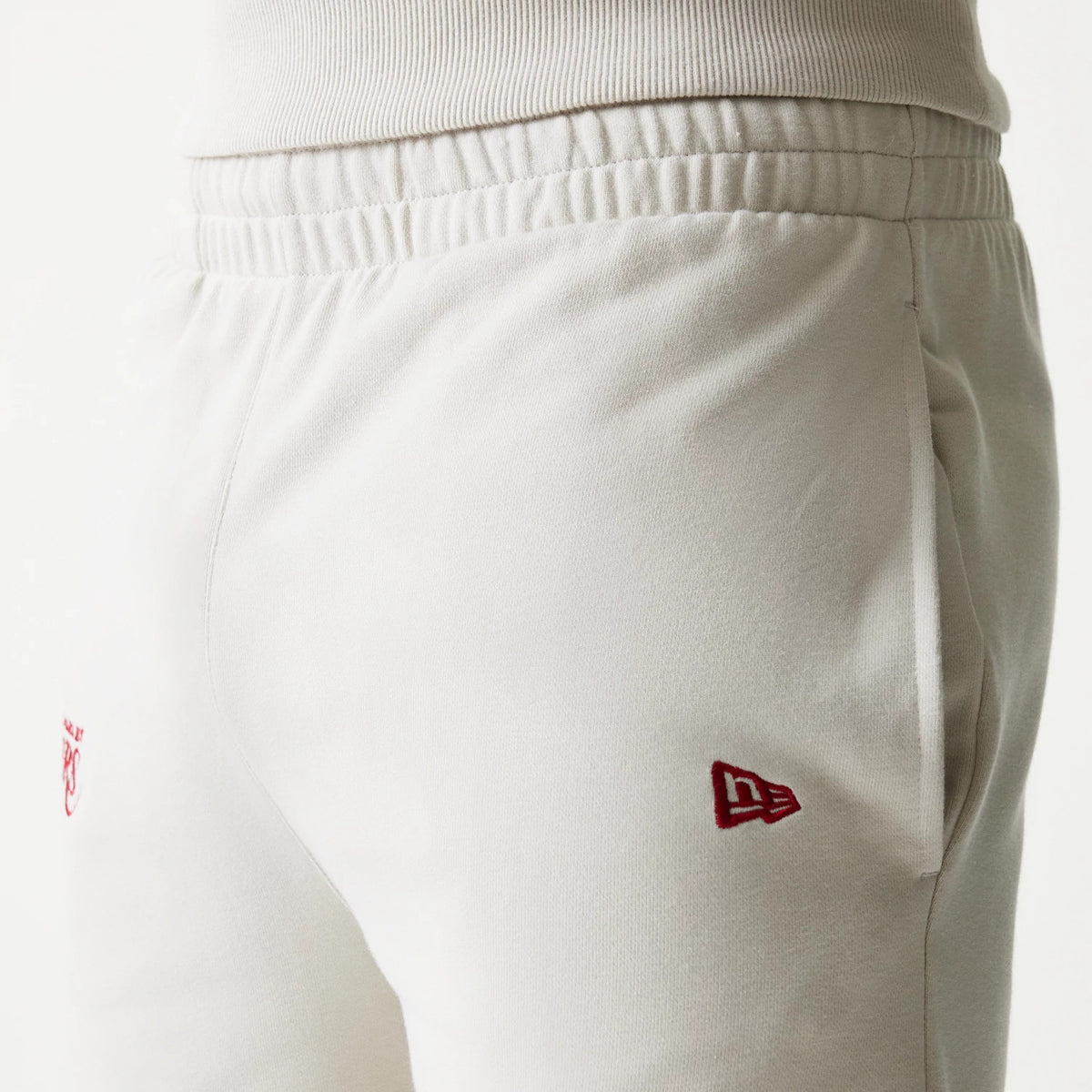 New Era LA Lakers NBA League Essential Light Beige Shorts, close-up side view showing an elastic waistband, a red embroidered New Era flag logo on the side near the pocket, and a small red LA Lakers logo slightly above the leg.