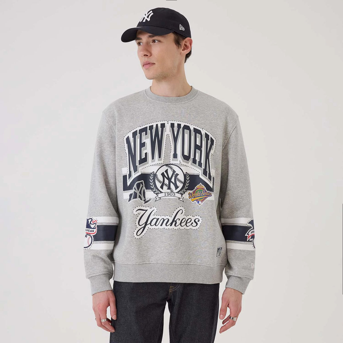 New Era New York Yankees MLB Sport Classic Grey Crew Neck Sweater, front view on a model with a matching cap, featuring large navy New York graphics, a Yankees logo, and World Series patches across the chest and sleeves.