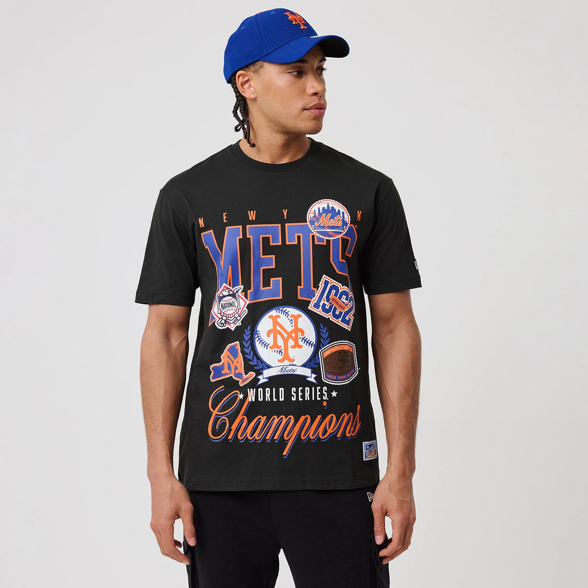 New Era New York Mets MLB Sport Classic Black T-Shirt, front view featuring Mets and Champions graphics in blue and orange across the chest, worn by a model with a matching blue cap.