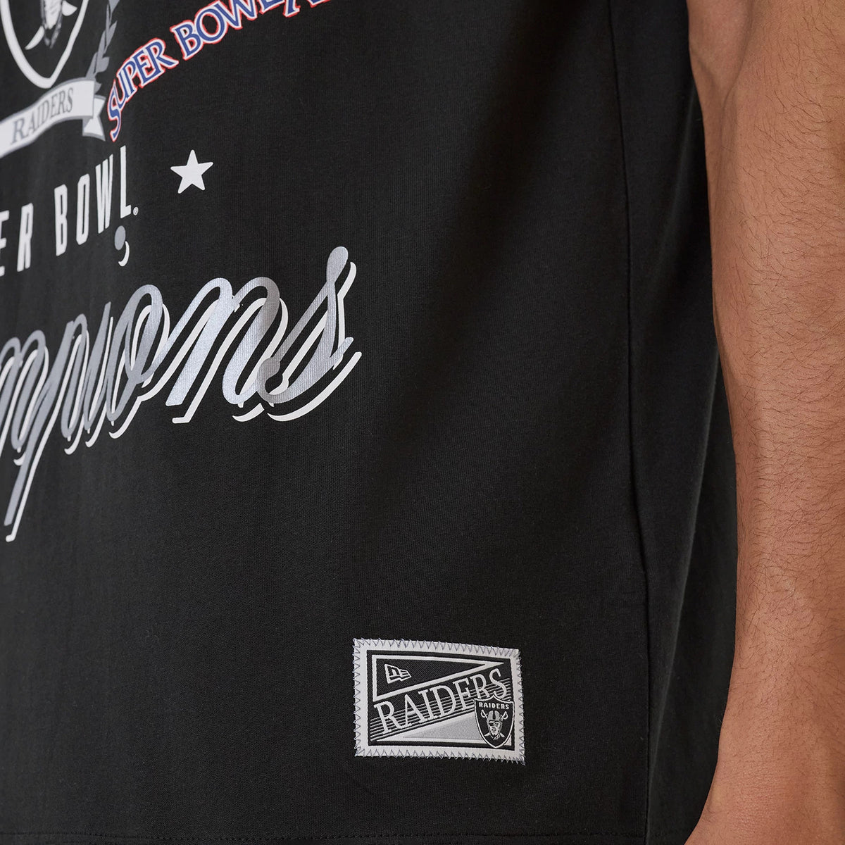 New Era Las Vegas Raiders NFL Sport Classic Black T-Shirt, front view showing white lettering spelling 'Champions' across the lower front, a printed Raiders patch near the hem, and detailed graphics of the team's achievements above.