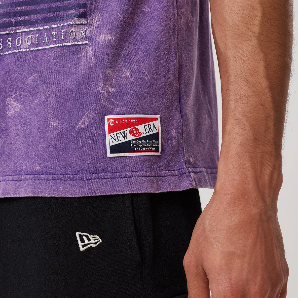 New Era LA Lakers NBA Purple T-Shirt, side view showing the lower portion with a vintage New Era logo patch near the hem, marbling throughout the purple fabric, and part of the print text above.