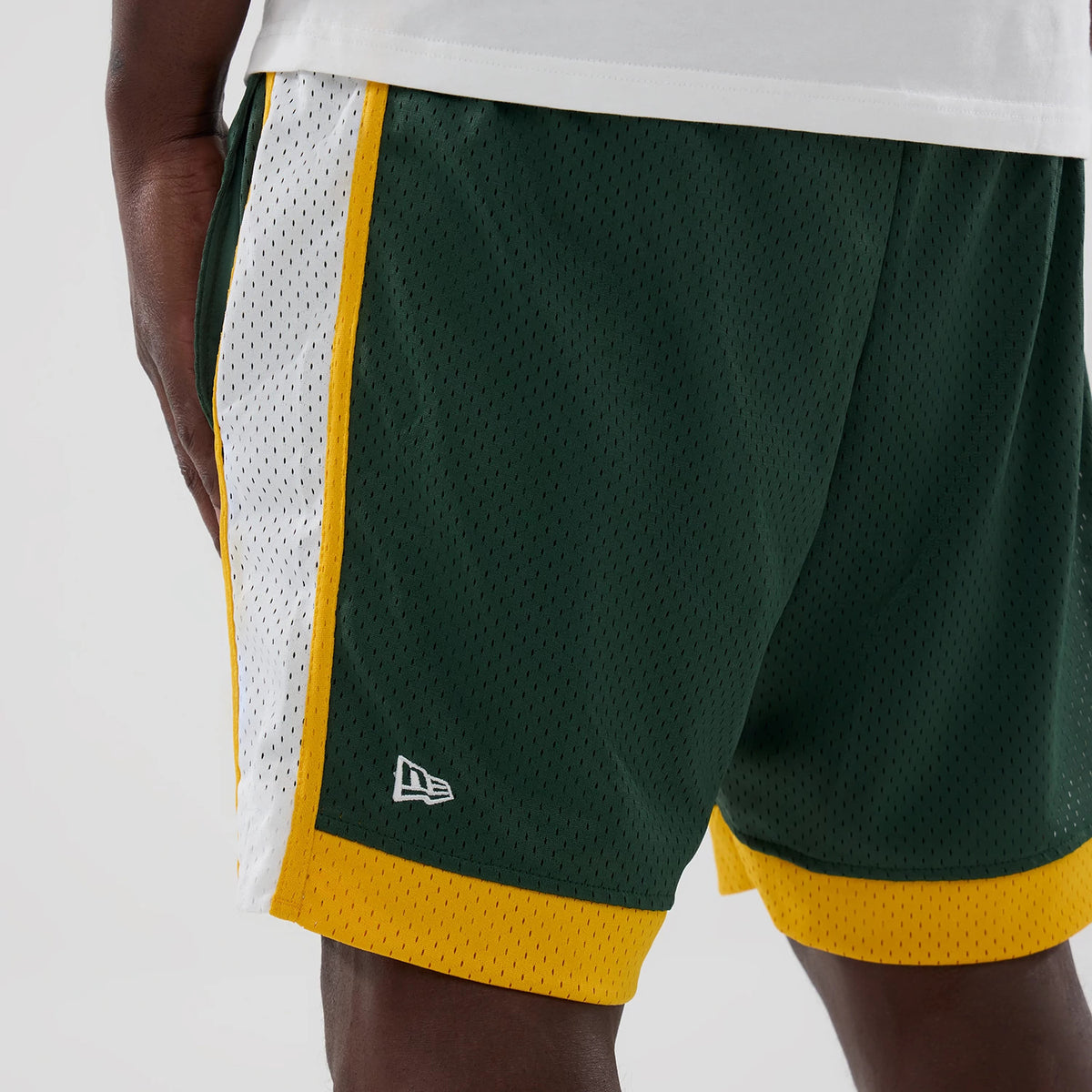 New Era Athletics MLB Dark Green Mesh Shorts, on-model front view showing a white panel with yellow trim on the sides, dark green mesh fabric, and an embroidered New Era logo above the yellow hem at the left leg.