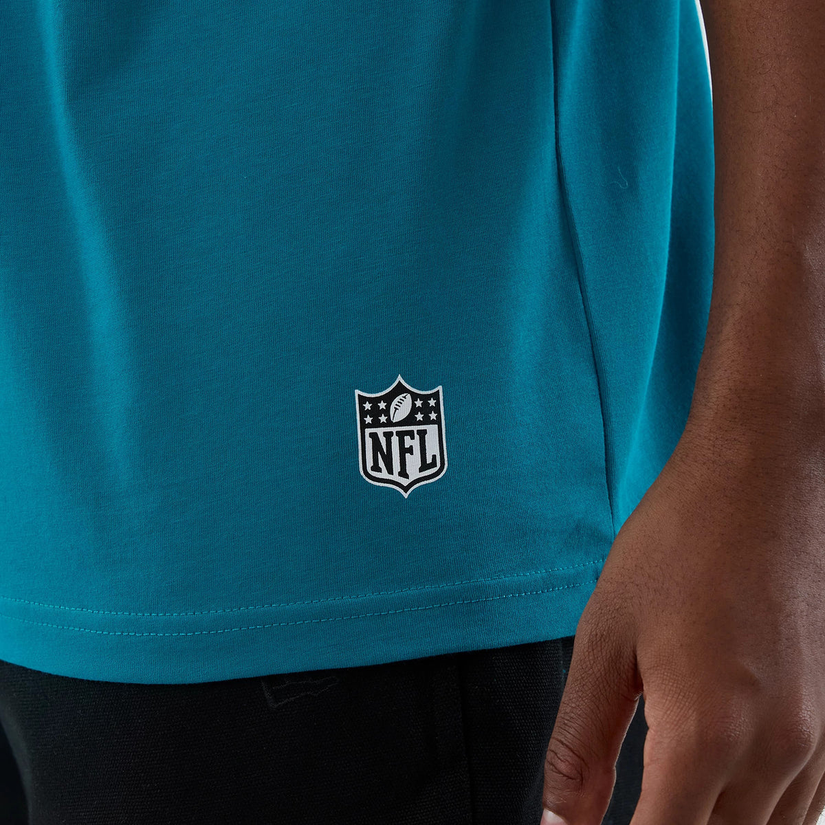 New Era Jacksonville Jaguars NFL International Series Games Turquoise Oversized T-Shirt, side view showing a white printed NFL logo near the hem, with the turquoise fabric falling loosely above black trousers.