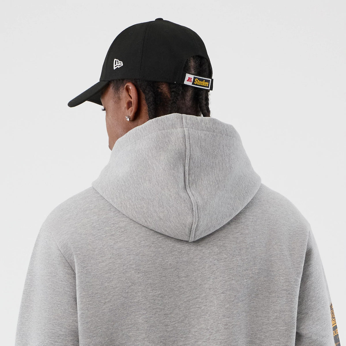 New Era Pittsburgh Steelers NFL International Series Games Grey Pullover Hoodie, back view on model showing the plain grey hood and shoulders, with a black cap featuring an embroidered New Era logo and a Steelers tab at the back.
