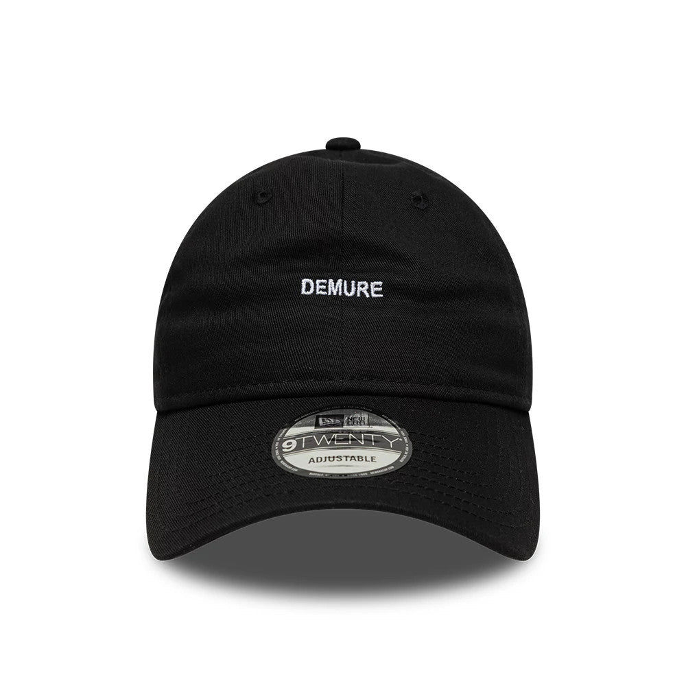 New Era Slogan Demure Black 9TWENTY Adjustable Cap, front view showing the white embroidered DEMURE text on the structured black crown, with a curved visor and 9TWENTY size sticker.