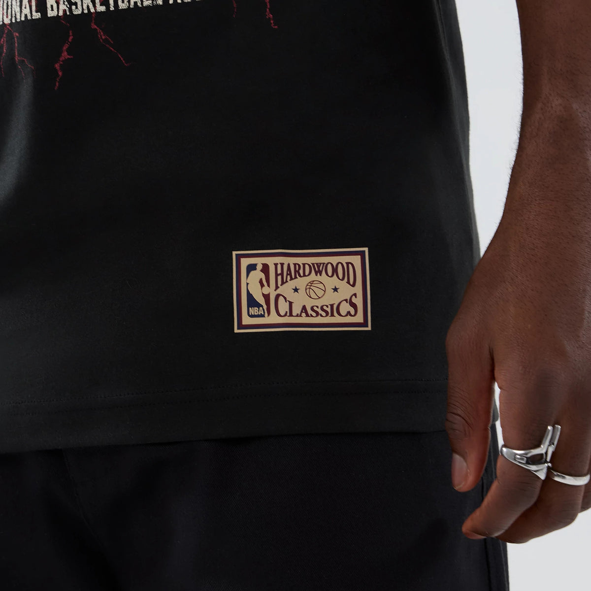 New Era Denver Nuggets NBA Hardwood Classic Black T-Shirt, side view showing a beige printed NBA Hardwood Classics patch near the hem, with parts of red lightning graphics above, styled on a model wearing black trousers.