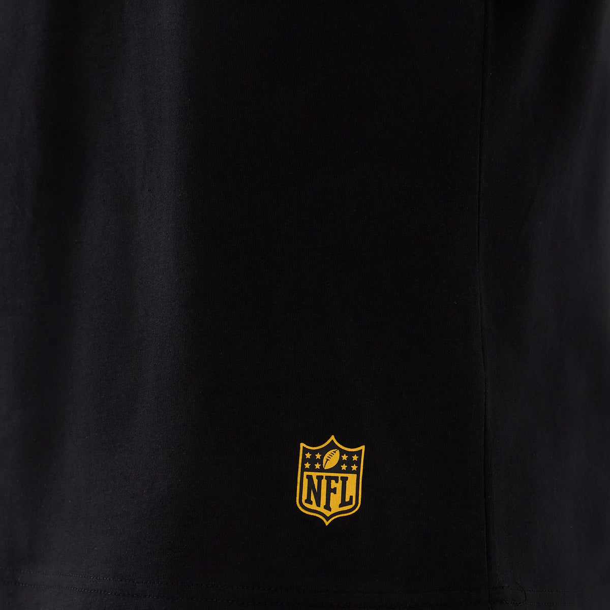 New Era Commanders NFL International Series Games Script 2025 Black Oversized T-Shirt, front view highlighting the small yellow embroidered NFL shield logo positioned near the bottom hem against the black fabric background.