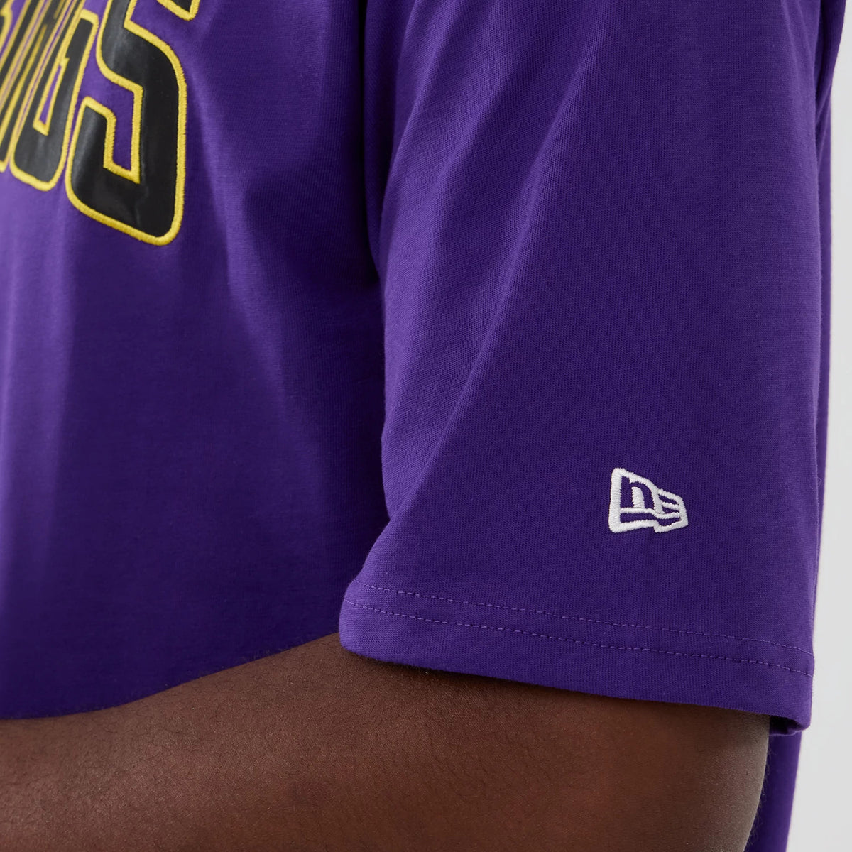 New Era Minnesota Vikings NFL International Series Games 2025 Purple Premium T-Shirt, side view showing the black and yellow Vikings lettering on the chest and the white embroidered New Era logo near the cuff on the short sleeve.
