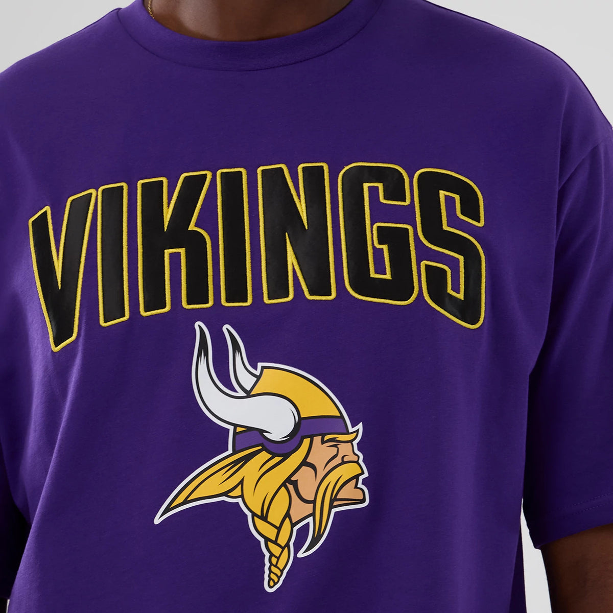 New Era Minnesota Vikings NFL International Series Games 2025 Purple Premium T-Shirt, front view on-model showing black and yellow outlined Vikings lettering across the chest with the team's Viking head logo printed below.