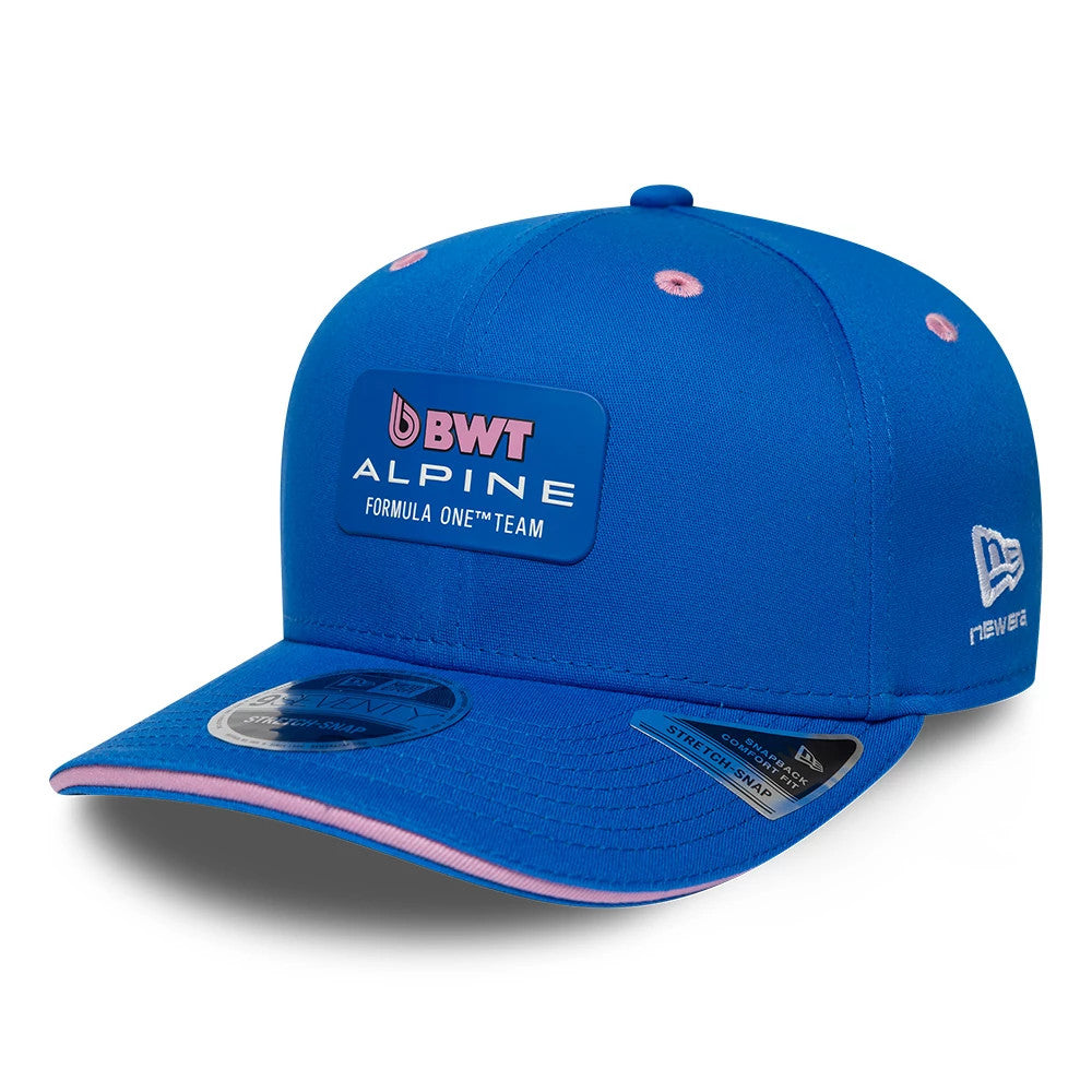 Alpine Racing Team Blue New Era 9SEVENTY Stretch Snap Adjustable Cap, three-quarter view with the BWT Alpine Formula One™ Team logo in pink and white on the blue front panel, pink undervisor and 9SEVENTY size sticker on the curved blue visor.