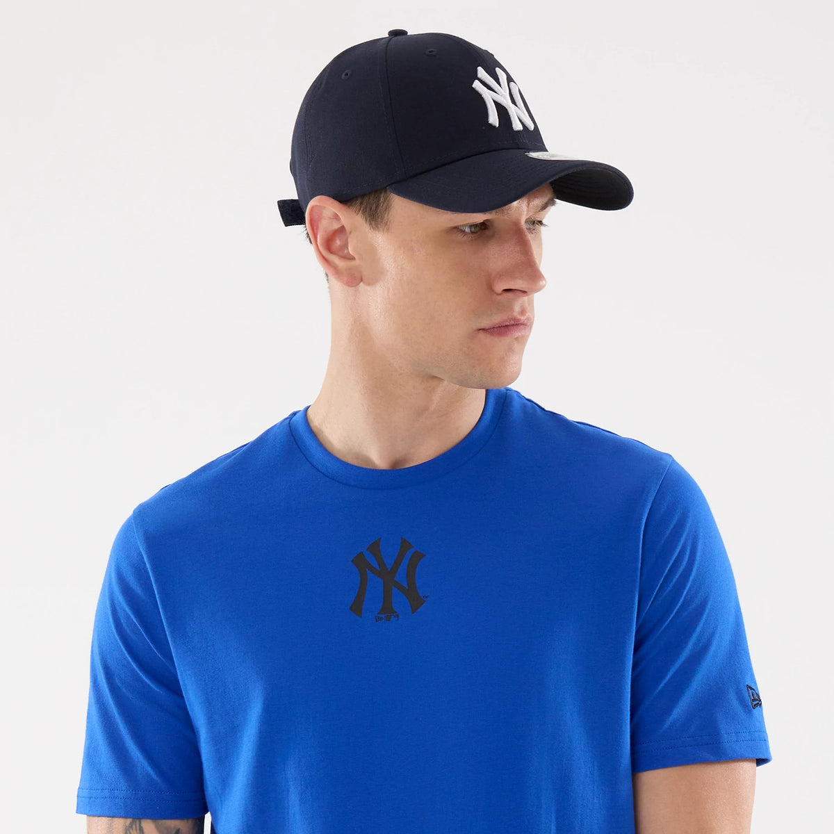 New Era New York Yankees MLB Blue T-Shirt, on-model view showing a royal blue T-shirt with a black printed New York Yankees logo centred on the chest, paired with a navy cap featuring a raised white NY logo and a small New Era logo on the sleeve.