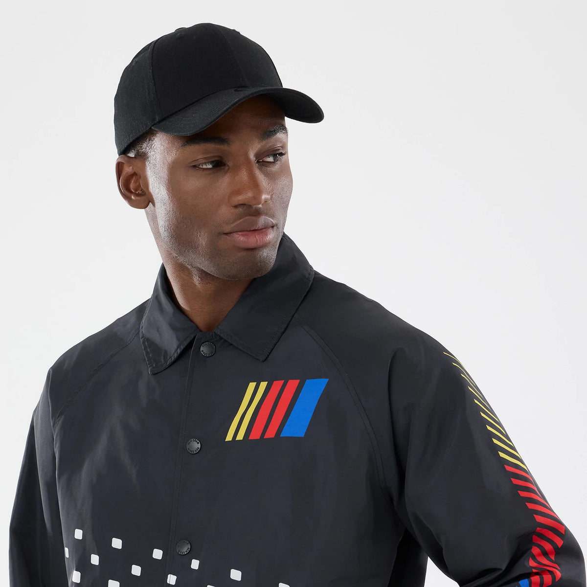 New Era Nascar Logo Black Jacket, front view on a model featuring a yellow, red, and blue embroidered Nascar logo on the chest, a series of white square prints at the bottom, and striped down the left sleeve.