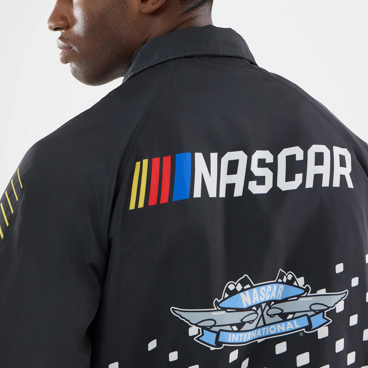 New Era Nascar Logo Black Jacket, back view showing white NASCAR lettering across the shoulders accompanied by colourful stripes, with a detailed printed Nascar International logo beneath, worn by a model for a fit.