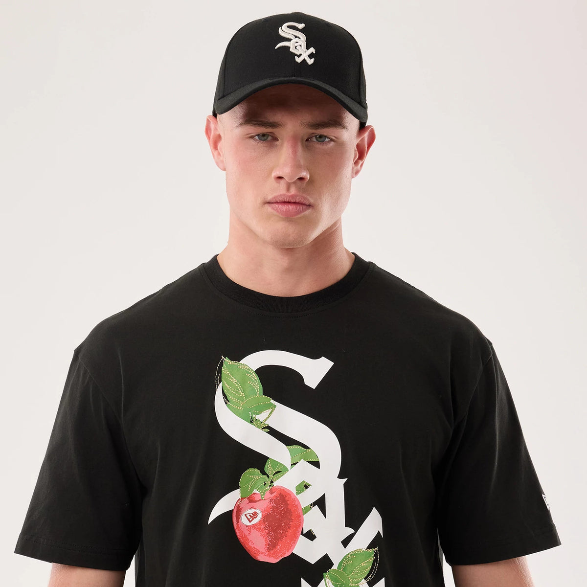 New Era Chicago White Sox MLB Life Graphic Black T-Shirt, front view on-model showing a large white Sox graphic intertwined with green leaves and a red apple on the chest, while the model wears a matching black cap with embroidered Sox logo.