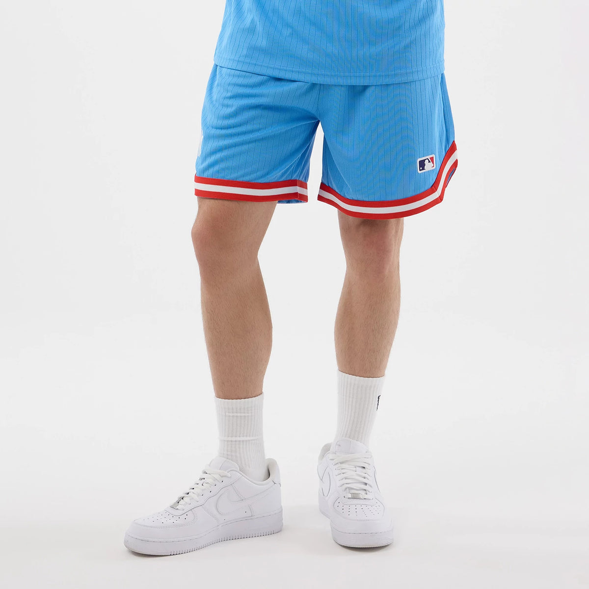 New Era New York Yankees MLB Arch Pastel Blue Oversized Shorts, on-model view showcasing light blue shorts with an embroidered MLB logo on the left leg, red and white striped hems at the bottom, paired with white trainers and socks.