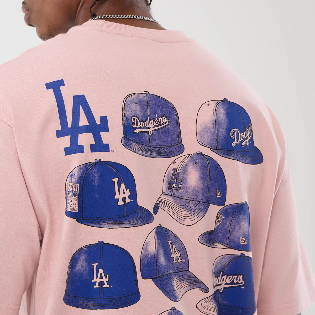 New Era LA Dodgers MLB Cap Graphic Pastel Pink Oversized T-Shirt, back view on a model showing multiple blue cap graphics featuring LA and Dodgers logos, with a large LA logo above, all set against the pastel pink fabric.