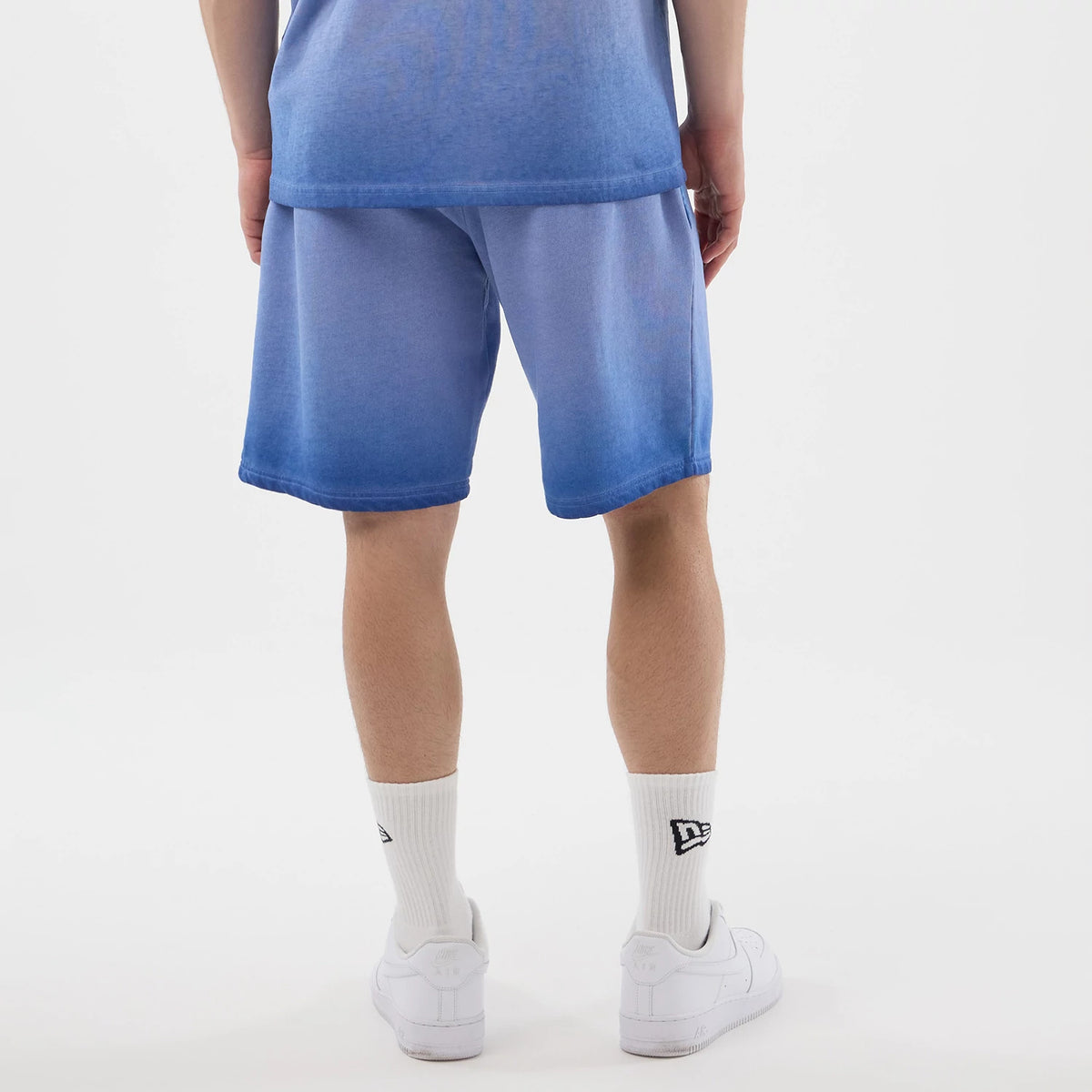 New Era LA Dodgers MLB Washed Logo Pastel Blue Shorts, back view on a model showing the gradient pastel blue shade of the shorts reaching just above the knees, styled with white socks featuring a black embroidered New Era logo and white trainers.