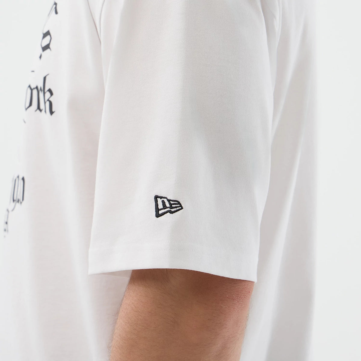 New Era Death Moth Graphic White Oversized T-Shirt, side view showcasing the short sleeve with an embroidered New Era logo near the hem, sections of gothic script lettering partially on the torso, and a relaxed fit on the model’s arm.