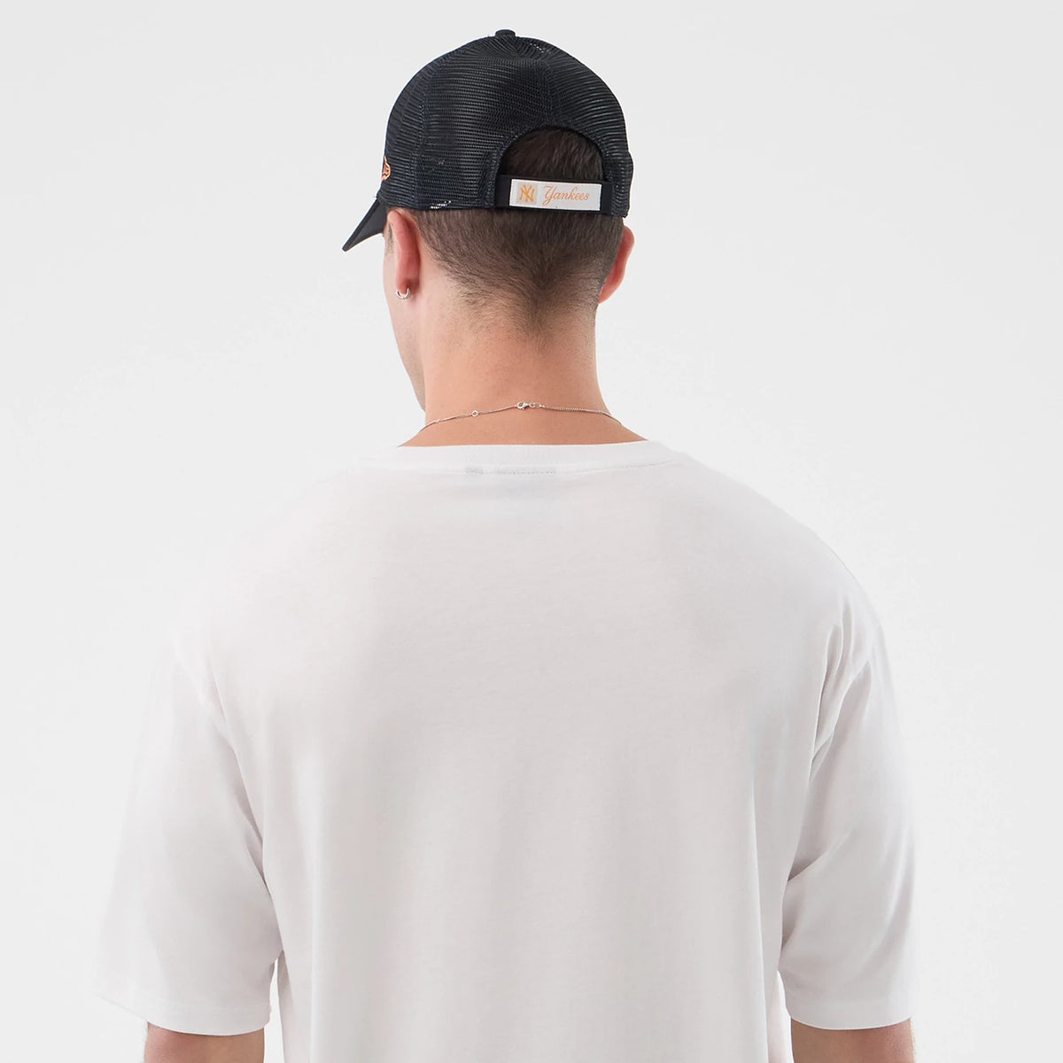 New Era Death Moth Graphic White Oversized T-Shirt, back view on-model showing the plain white fabric extending over the shoulders, loosely fitted sleeves and a rounded neckline; styled with a dark cap from the rear.
