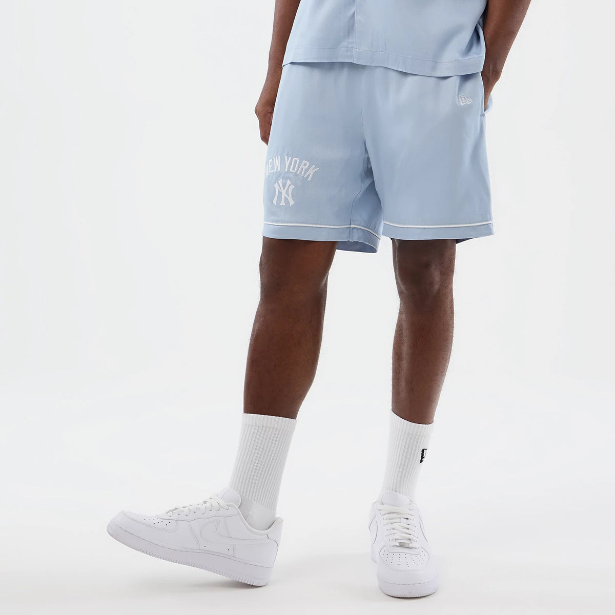 New Era New York Yankees MLB Woven Pastel Blue Shorts, front view on a model showing the light blue colour with white piping along the hem, an embroidered New Era logo near the pocket and New York lettering above the Yankees logo on the left leg.