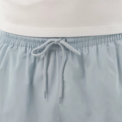 New Era Pastel Blue Swimming Shorts, front view showing the light blue fabric with an elasticated waistband, a drawstring tied in the centre, and the top edge of the paired white shirt just above the shorts.