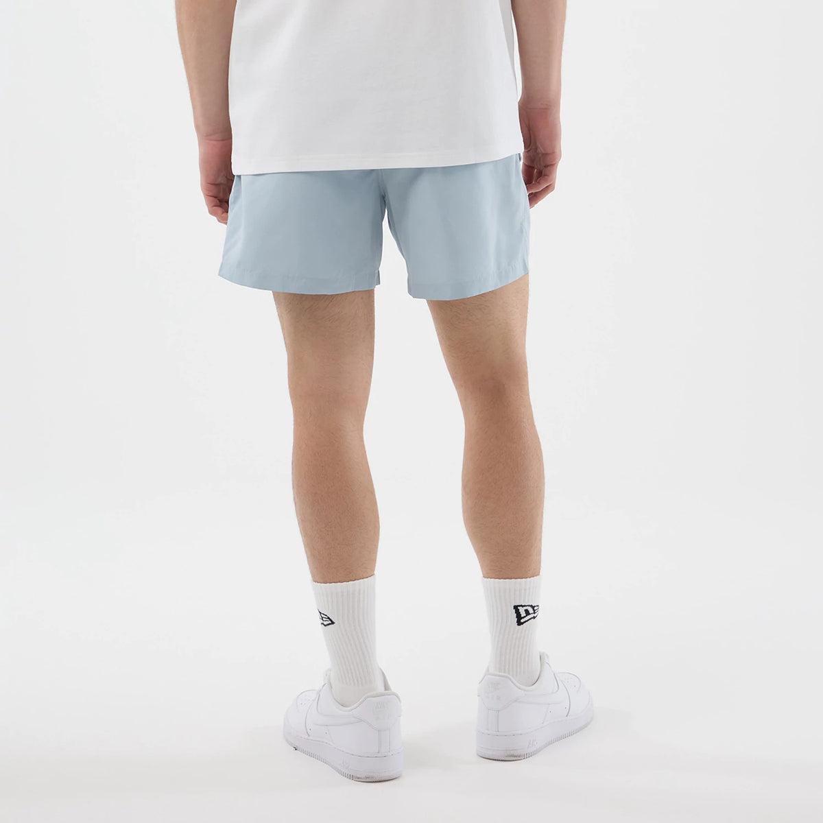 New Era Pastel Blue Swimming Shorts, back view on a model wearing white socks and trainers, showing the clean lines of the pastel blue shorts contrasting against a plain white shirt, no logos or patterns present.