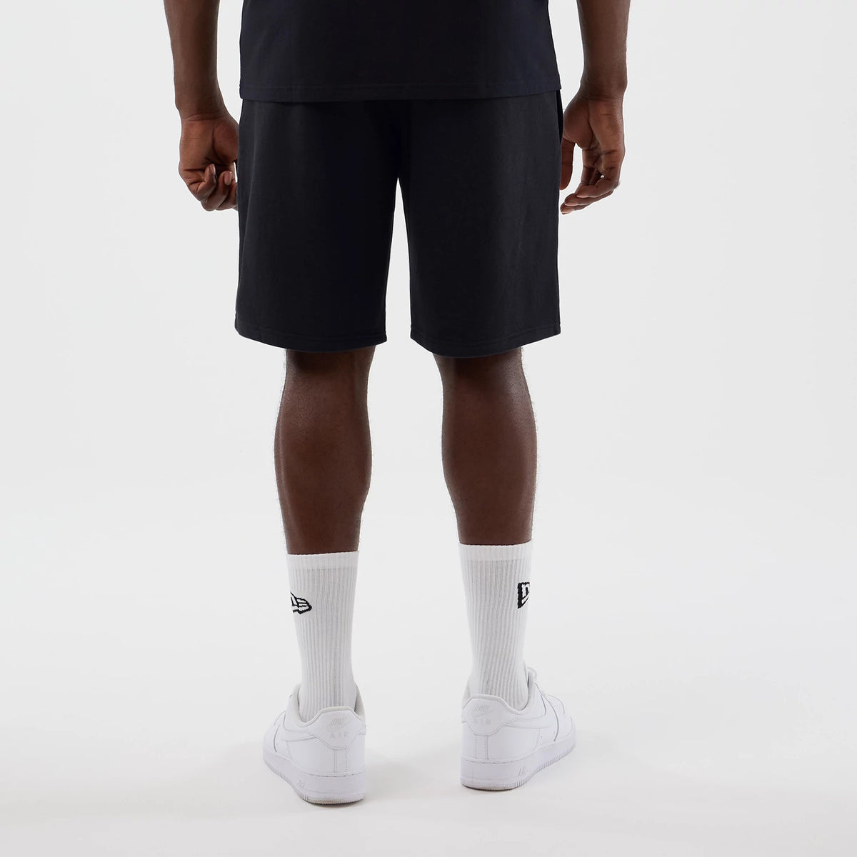 New Era LA Lakers NBA Core Plus Navy Shorts, back view on a model with a plain black T-shirt, showcasing the solid navy colour and mid-length design, complemented by white socks featuring an embroidered New Era logo and plain white trainers.