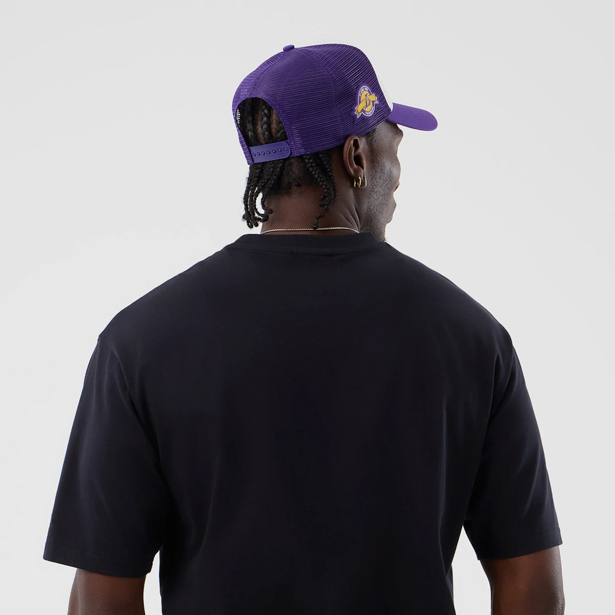 New Era LA Lakers NBA Core Plus Navy Oversized T-Shirt, back view on a model paired with a purple cap, showing the plain navy fabric extending over the shoulders and partway down the short sleeves.