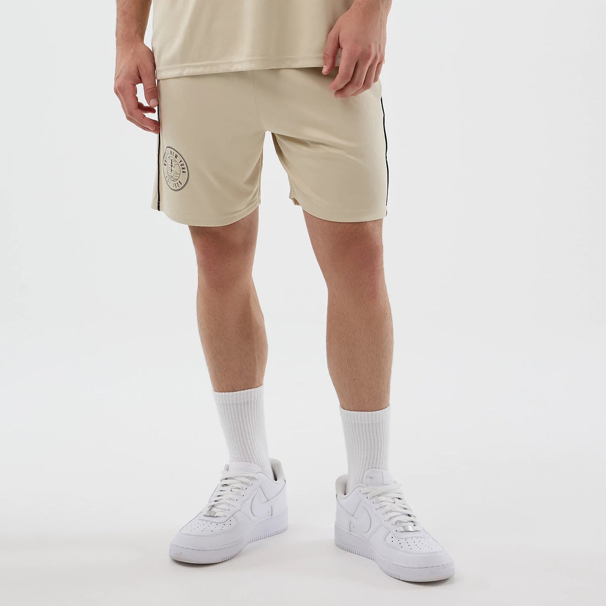 New Era Football Light Beige Shorts, front view on-model showing an embroidered New Era logo on the left leg, with a drawstring waist and a contrasting black stripe running down the side seam, styled with white trainers and socks.