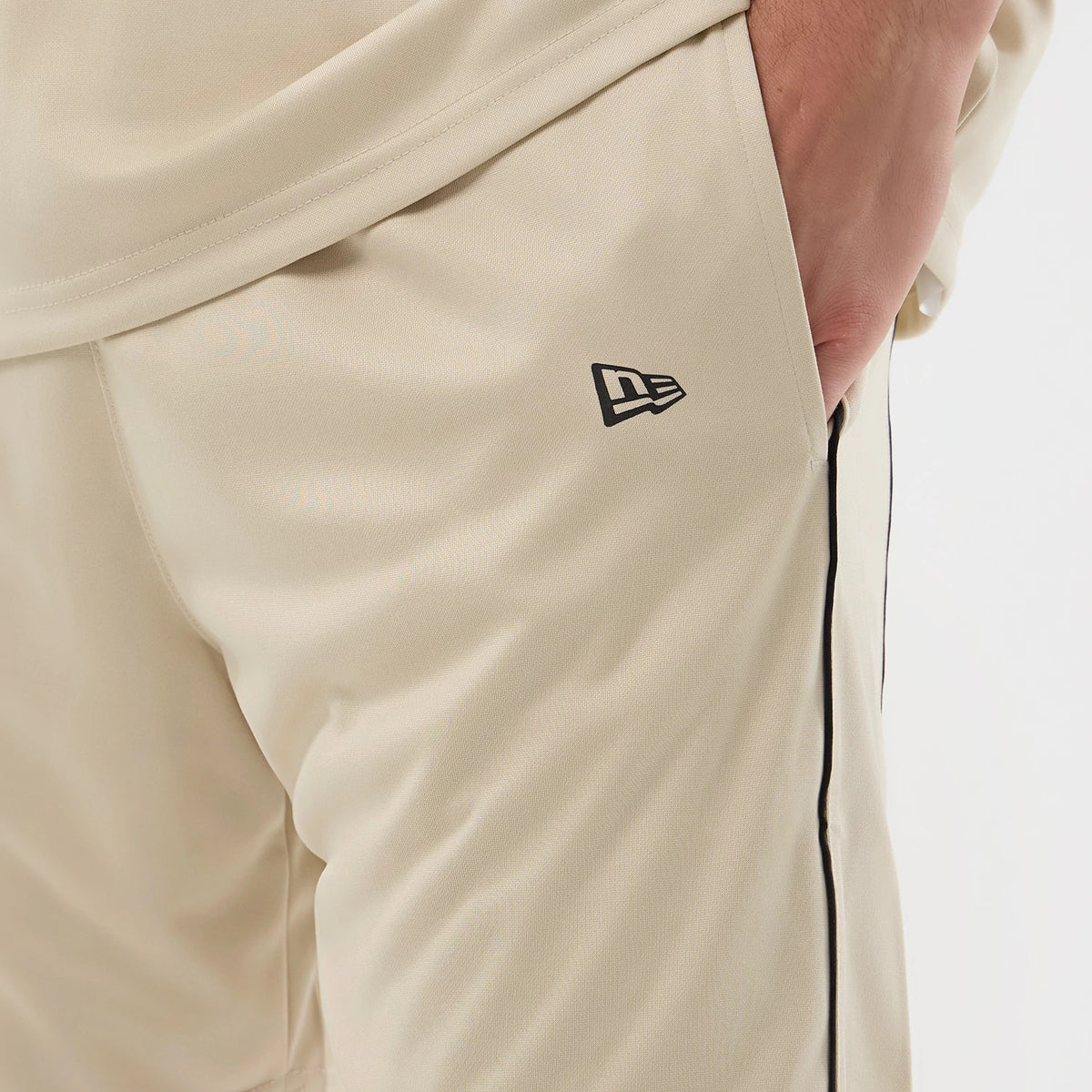 New Era Football Light Beige Shorts, three-quarter view with black embroidered New Era logo near the left pocket, black piping running along the side seam, and a view of an elastic waistband at the top.