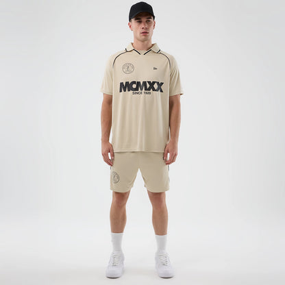 New Era Football Light Beige Shorts, front view on a model styled with a matching shirt and black cap, featuring an embroidered circular logo on the left leg, an elastic waistband, and a relaxed knee-length fit.