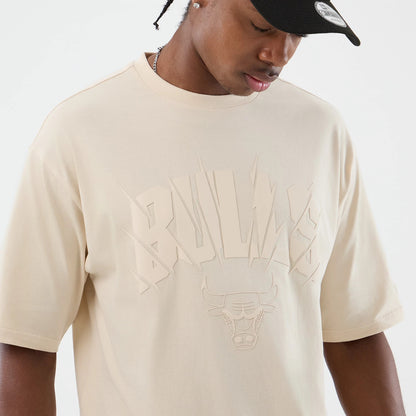 New Era Chicago Bulls NBA Core Plus Light Beige Oversized T-Shirt, front view on-model displaying the large embossed Bulls graphic across the chest, short sleeves, and a relaxed fit, paired with a black cap featuring a circular emblem on the front.