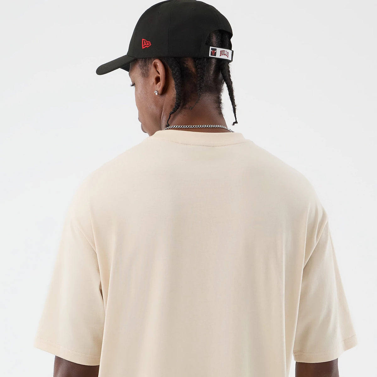 New Era Chicago Bulls NBA Core Plus Light Beige Oversized T-Shirt, back view on-model showcasing the plain light beige oversized design with short sleeves, styled with a black cap featuring embroidered red Bulls branding above the adjustable strap.