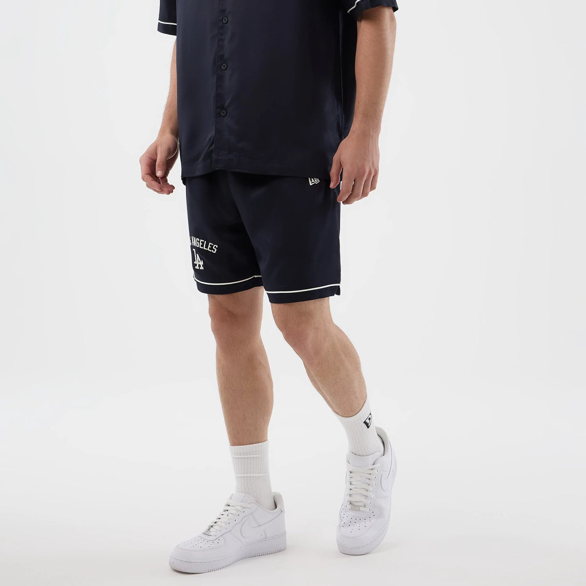 New Era LA Dodgers MLB Woven Navy Shorts, on-model view showing the navy shorts with white embroidered Los Angeles Dodgers lettering and logo on the left leg, contrasted by white trim at the hem and a New Era logo above.