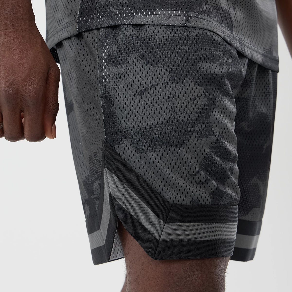 New Era All Over Print Mesh Black Camo Shorts, side view on-model showing the grey and black camo pattern along the fabric, a wide hem with black and grey stripes, and a slight slit at the side for added style.