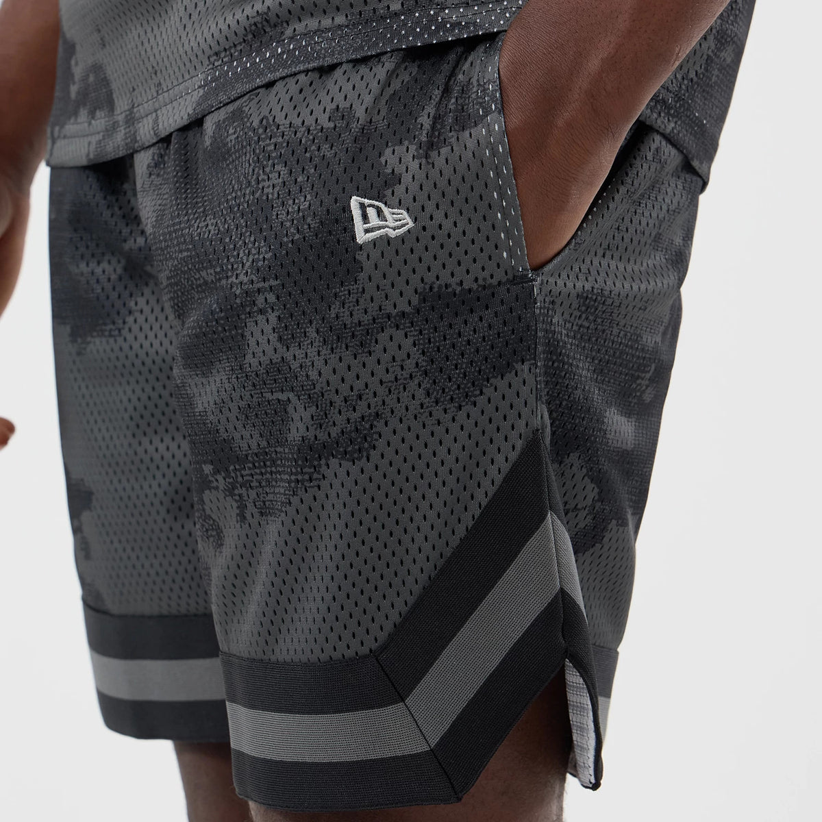New Era All Over Print Mesh Black Camo Shorts, side view showing the white embroidered New Era logo on the upper thigh, detailed black camo pattern across the mesh fabric, and wide horizontal stripes at the edge above the hem.