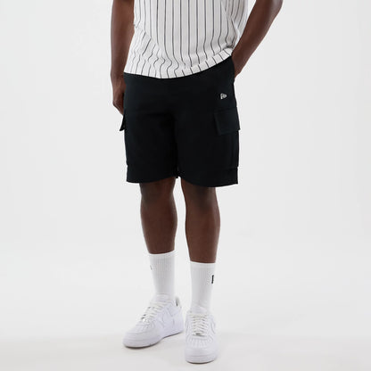 New Era Black Cargo Shorts, front view on a model styled with a pinstriped shirt and white trainers, displaying spacious cargo pockets on each side and a distinctive embroidered New Era logo on the left front pocket.