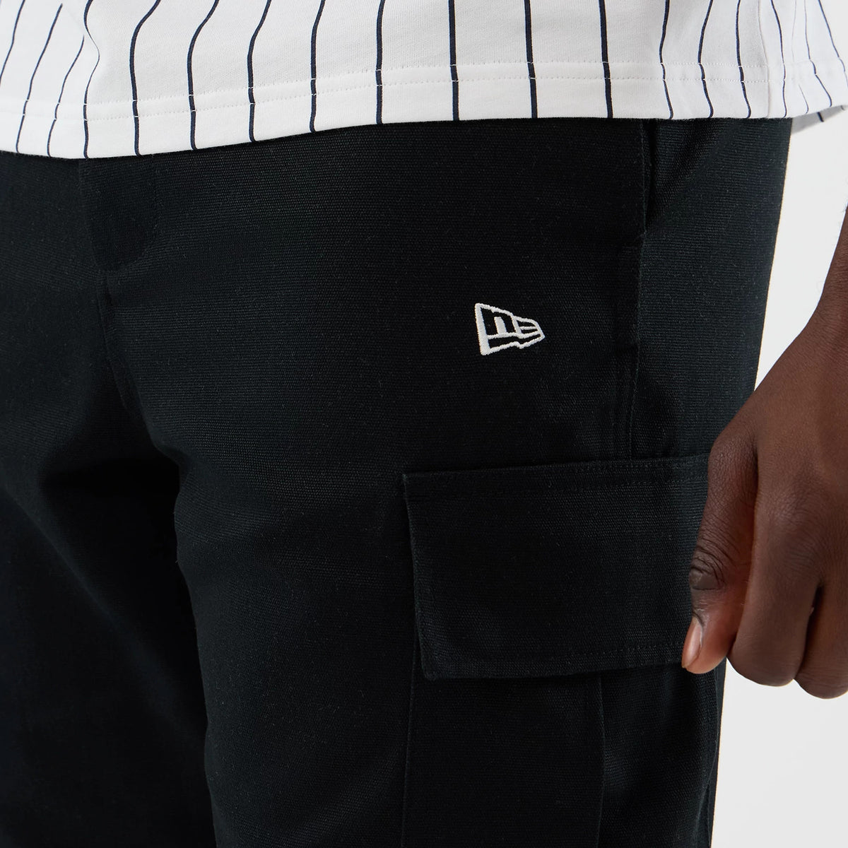 New Era Black Cargo Shorts, side view showing an embroidered white New Era logo on the pocket, a black cargo pocket with a flap closure, and a white pinstripe shirt partially above the waistband.