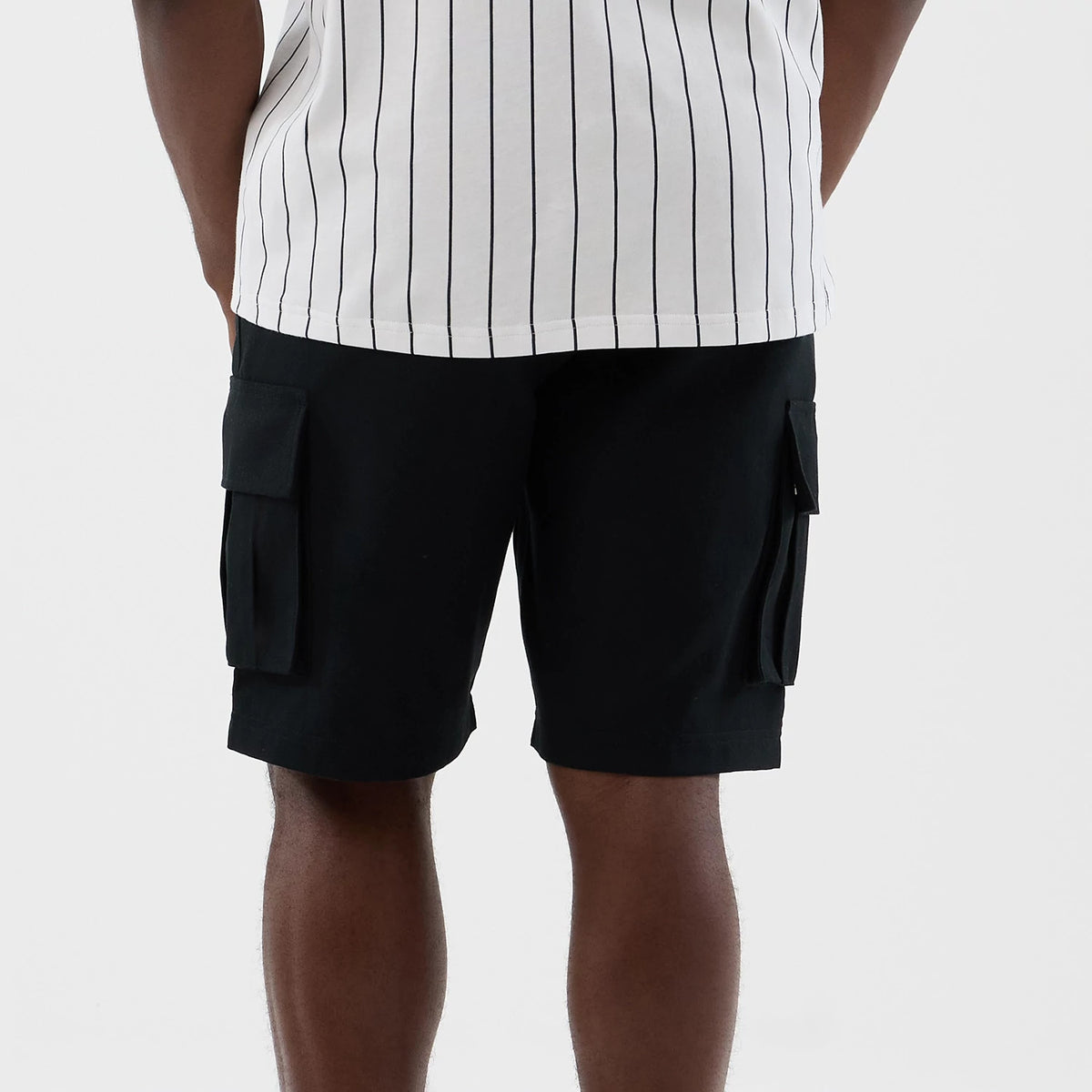New Era Black Cargo Shorts, back view on a model styled with a white pinstripe shirt, showing two large cargo pockets on each side of the shorts and a clean hemline reaching just above the knee.