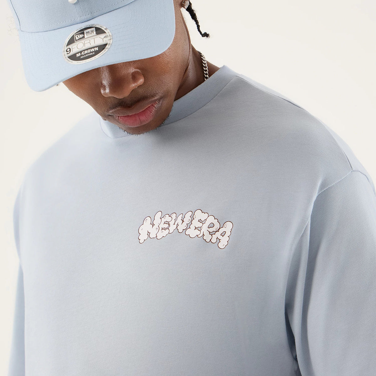 New Era Fun Block Party Graphic Pastel Blue Oversized T-Shirt, front view on-model showing a cloud-themed printed New Era logo on the chest, styled with a matching pastel blue new era cap featuring an embroidered logo on the front.