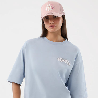 New Era Fun Block Party Graphic Pastel Blue Oversized T-Shirt, front view on-model featuring a playful cloud-like printed New Era logo on the left chest, short sleeves, and a relaxed fit, paired with a pastel pink cap.